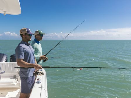 Two men stand on a boat fishing, casting lines into calm blue-green water beneath a clear sky.