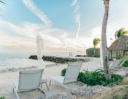 A tropical beach with white sand, lounge chairs, a closed umbrella, palm trees, a calm sea, and thatched huts on a sunny day.
