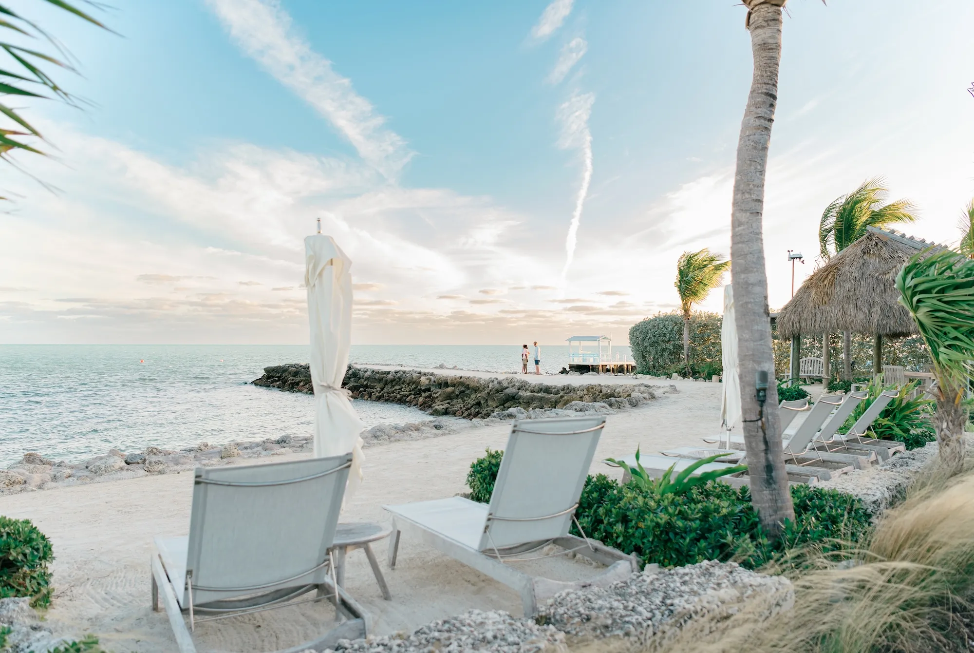 A tropical beach with white sand, lounge chairs, a closed umbrella, palm trees, a calm sea, and thatched huts on a sunny day.