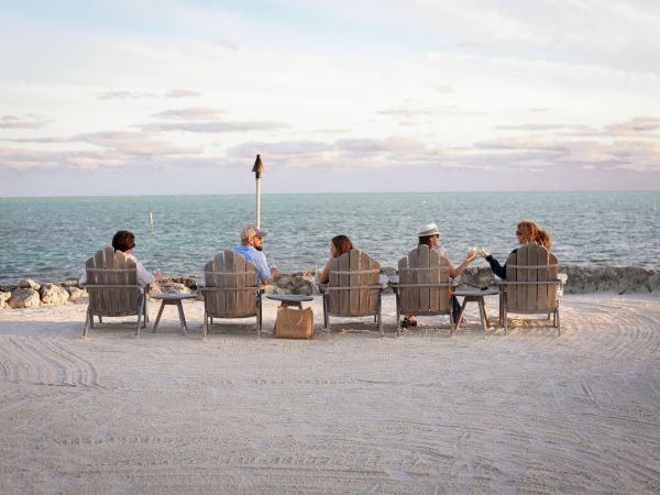 A group of friends sits in beach chairs facing the sea, sharing a quiet moment as a lone umbrella stands upright in the sand.