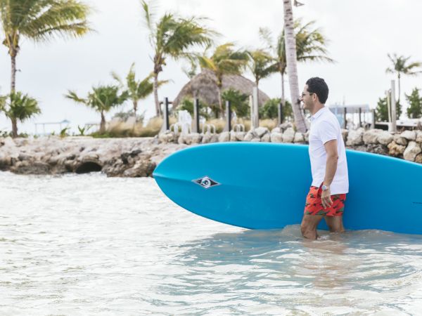 A man stands knee-deep in clear water with a bright blue surfboard, tropical palm trees, and a thatched-roof hut in the background.