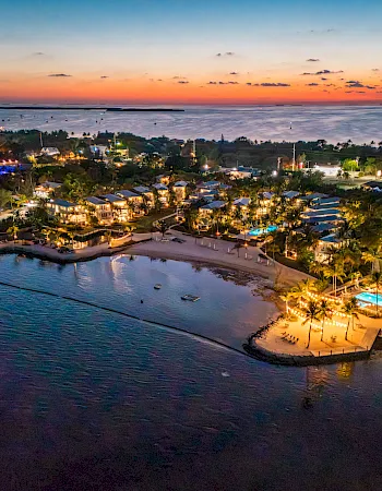 A beachfront resort glows at dusk with palm-lined villas, a sandy cove, boats, and calm ocean under a colorful sunset.