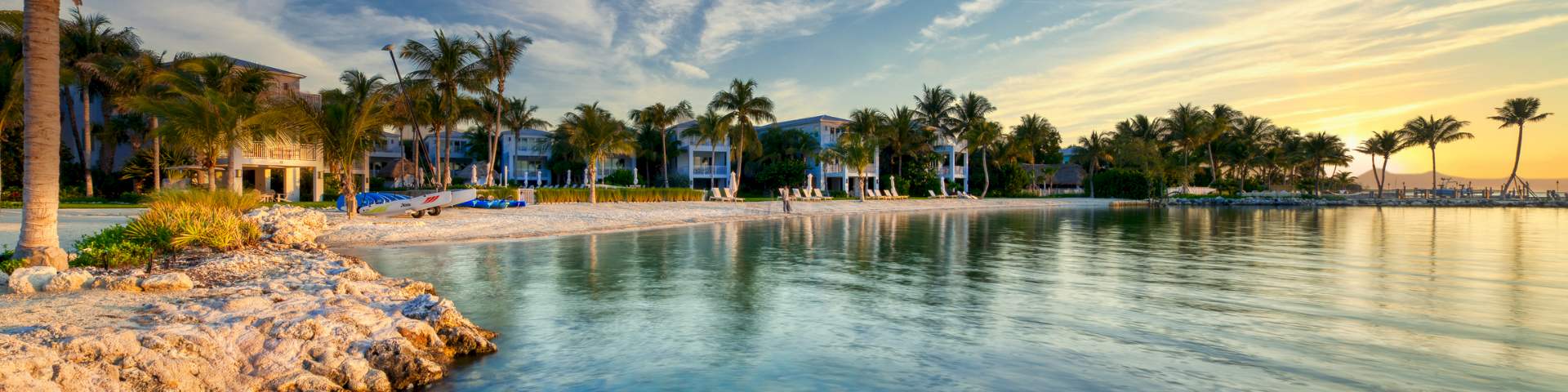 Tropical beachfront at sunset with palm trees and a quiet resort bay, calm water reflecting the colorful sky.