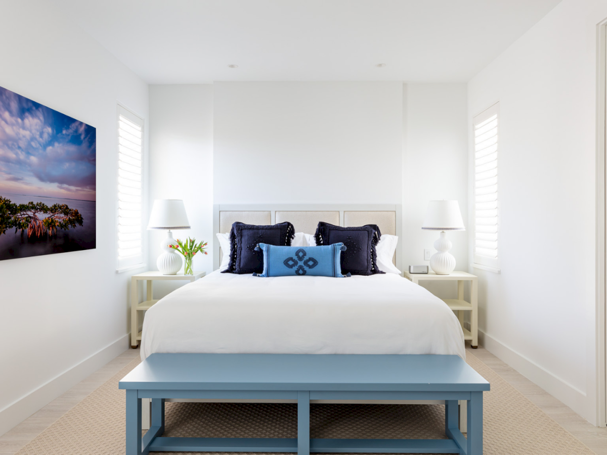 Bright modern bedroom with bed, pillows, nightstands, wall art, blue bench, and an open doorway to a tiled bathroom.