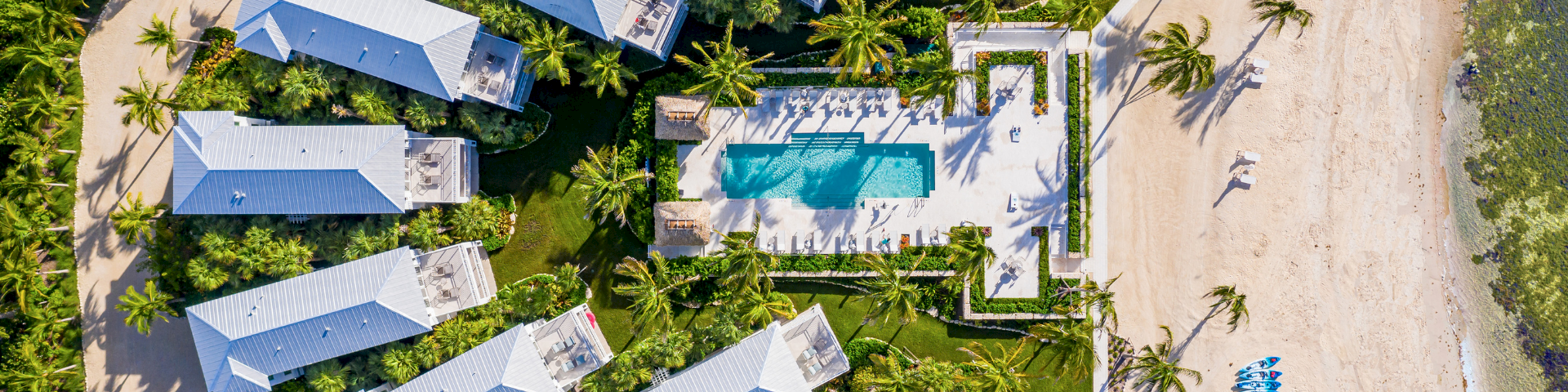 Aerial view of a tropical resort: solar-roof villas, lush palms, and a turquoise pool beside a sandy beach path.
