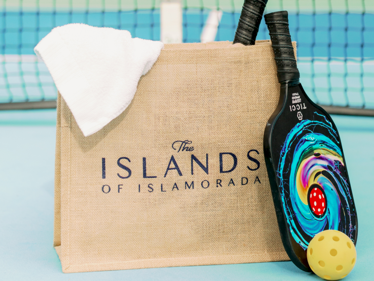 A burlap &ldquo;The Islands of Isla&hellip;&rdquo; bag by a pool, with a pickleball paddle, yellow ball, and a white towel.