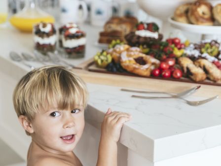 A little child stands by a kitchen food spread&mdash;bread, berries, and fruit&mdash;while looking toward the camera.