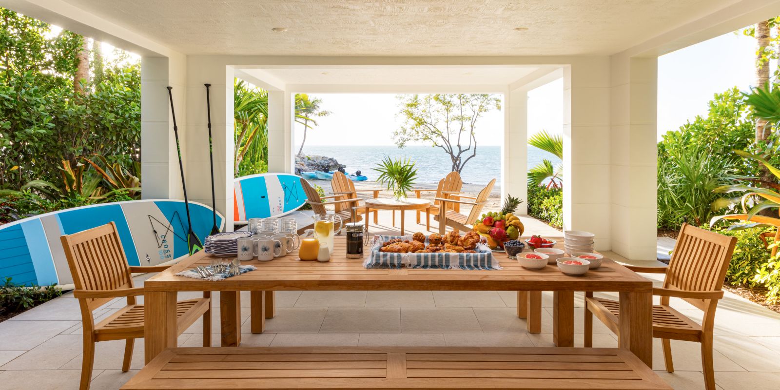 An oceanfront patio breakfast table with pastries, fruit, drinks, and surfboards by a lush garden and open sea view.