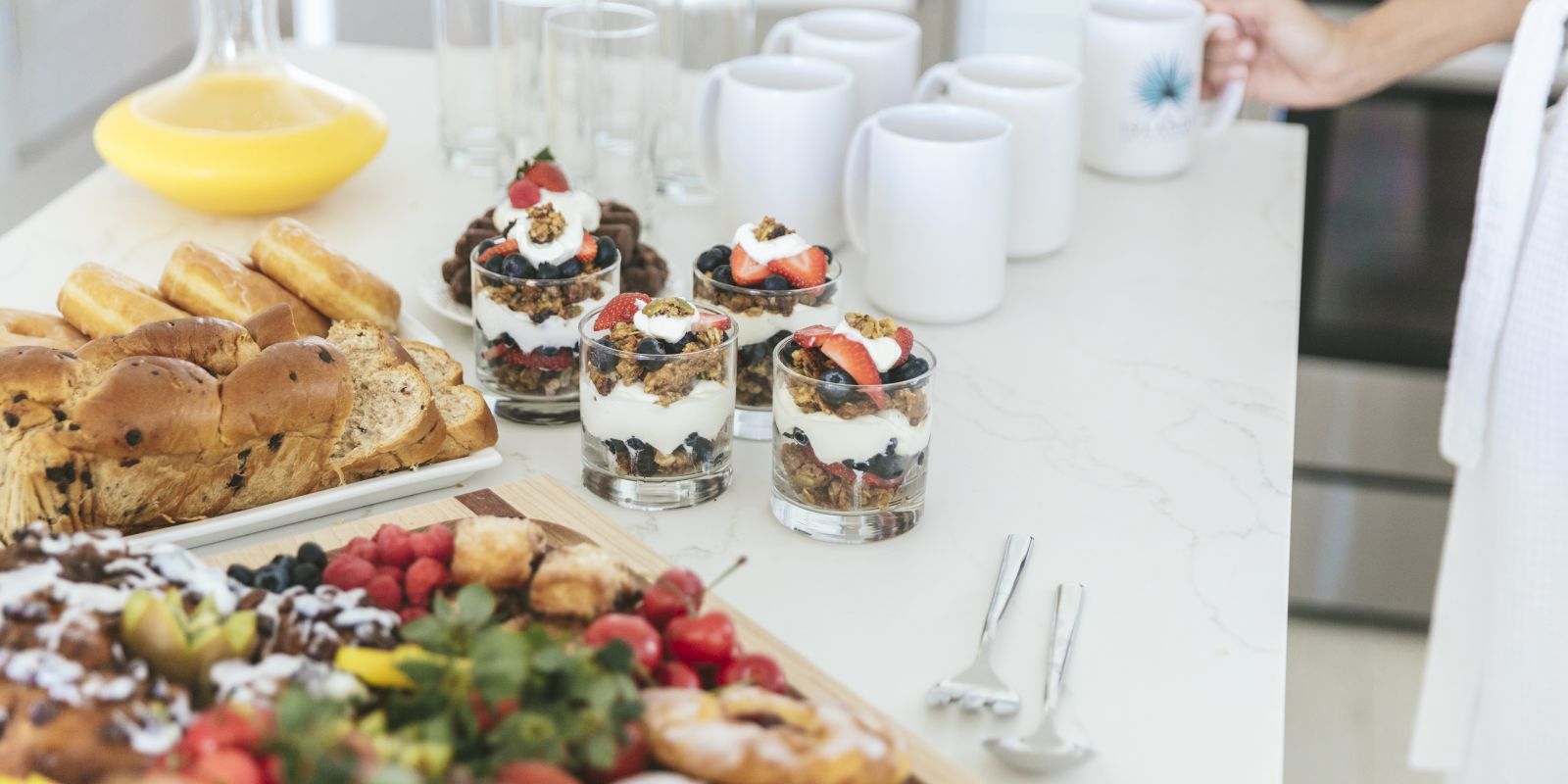A brunch spread: glasses of yogurt parfaits with berries and granola, bread rolls, and someone pouring a drink at a table.