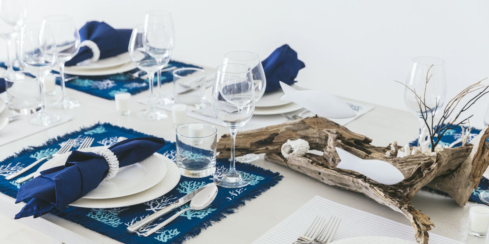 A festive blue-themed table setting with wine glasses, white plates, candles, cutlery, and a centerpiece of driftwood and ornaments.