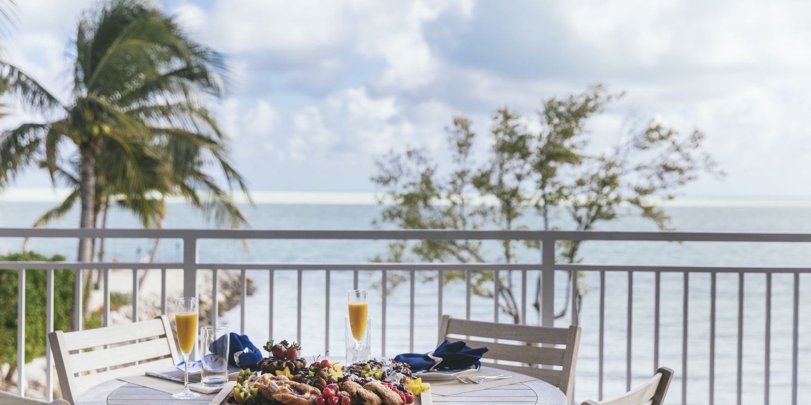A seaside patio breakfast table with champagne, fruit, and flowers, with palm trees and ocean waves under a blue sky.