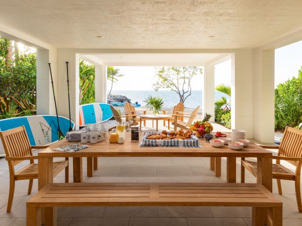 A sunny beachfront patio breakfast spread with fruit and pastries, amid tropical greenery and surfboards by the ocean.