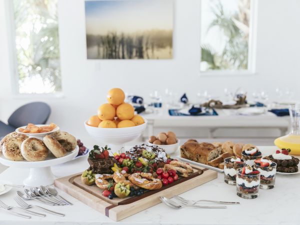 A bright breakfast buffet table with fresh fruit, pastries/bagels, waffles, yogurt and juice in a modern room setup.