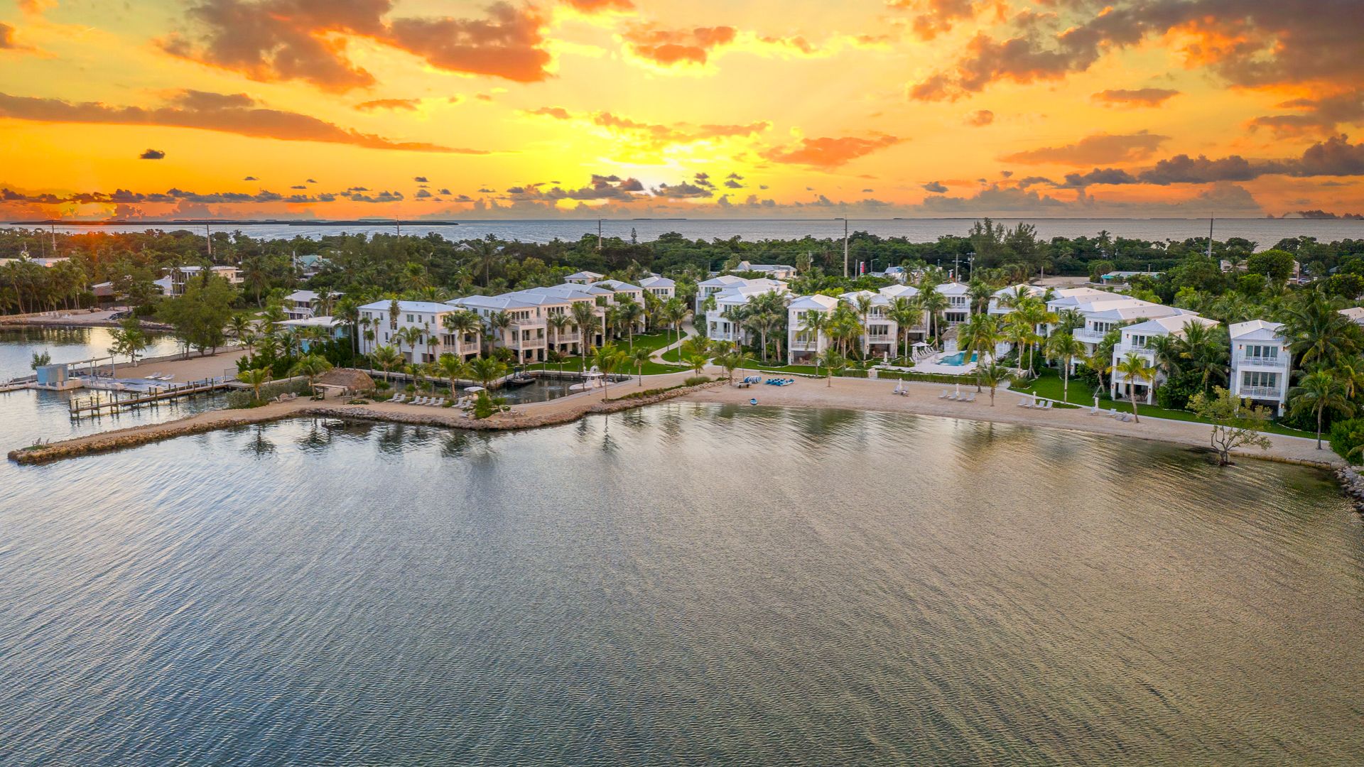 A beachfront resort with white villas, palms, docks, and calm sea under a dramatic sunset sky.