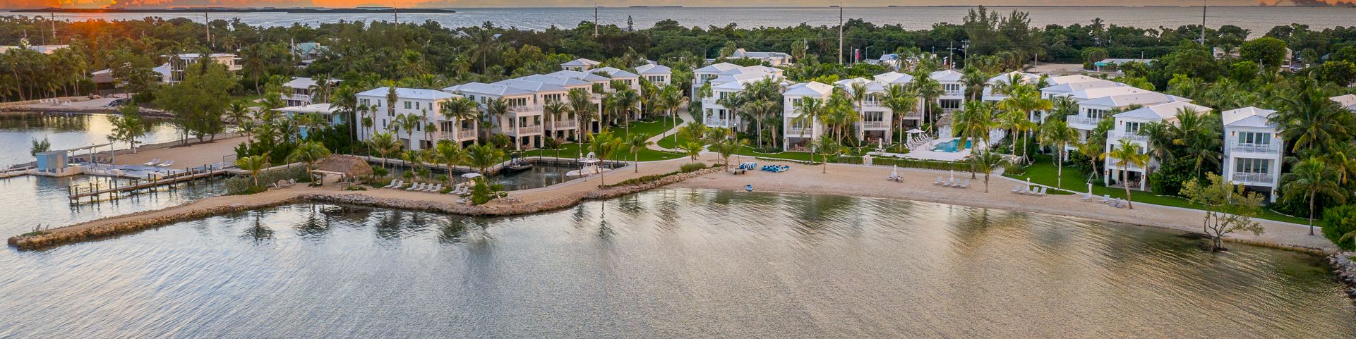 A beachfront resort with white villas, palms, docks, and calm sea under a dramatic sunset sky.