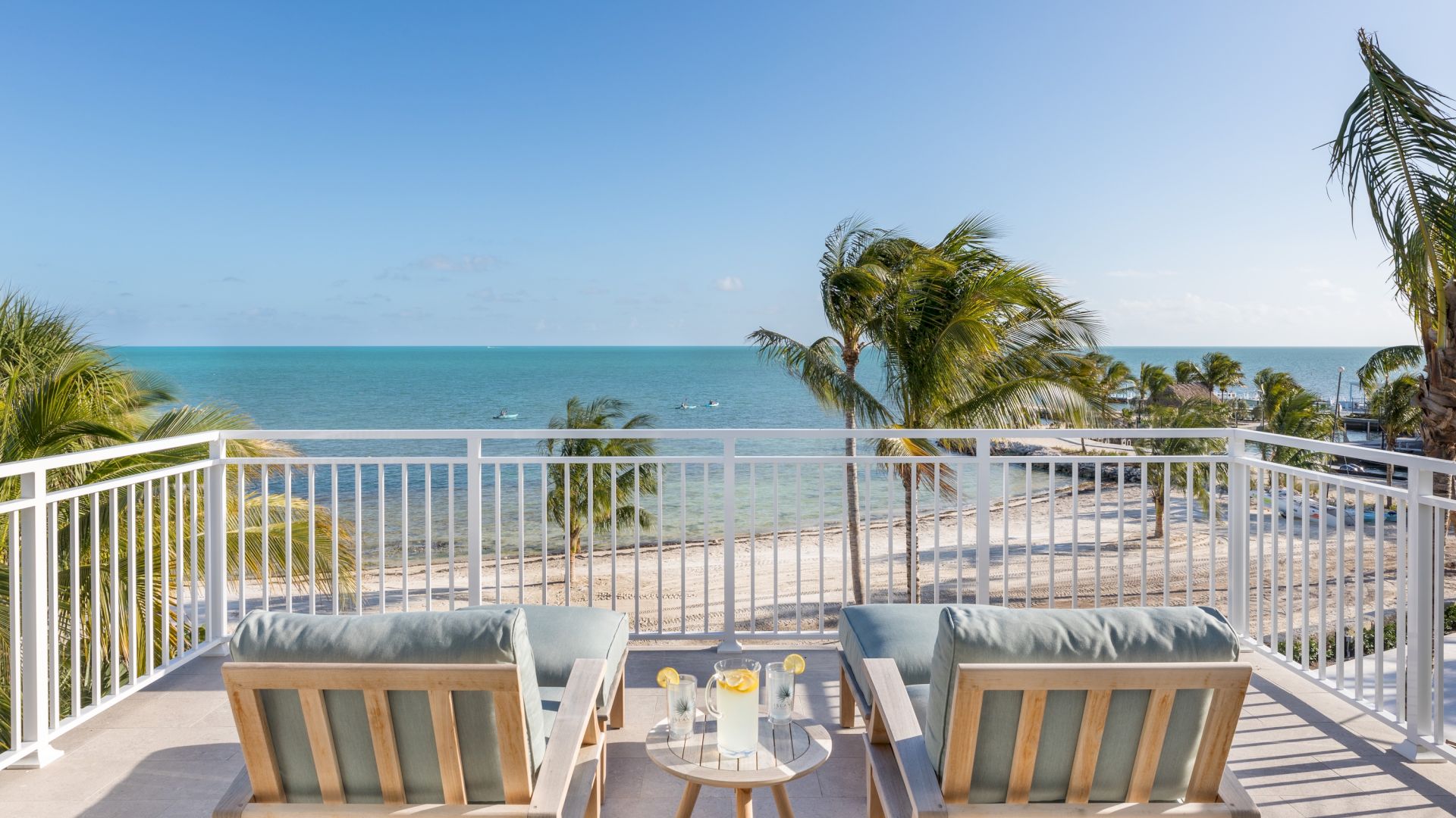 A sunny oceanfront balcony with palm trees, white railing, and two lounge chairs facing the turquoise sea.