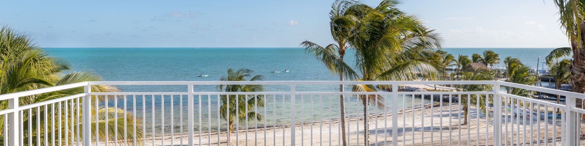 A sunny oceanfront balcony with palm trees, white railing, and two lounge chairs facing the turquoise sea.