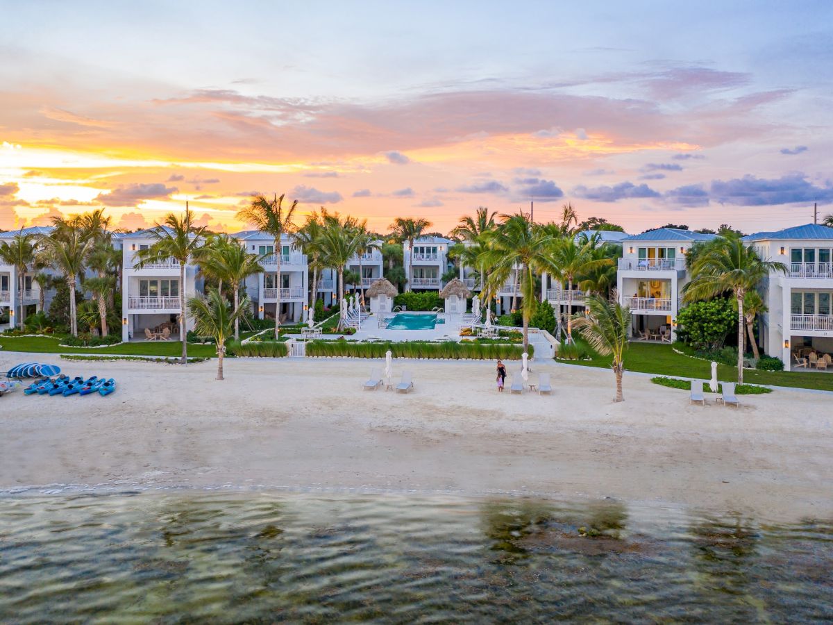 A tropical beachfront resort with white villas, palm trees, a central pool, and a glowing sunset sky.
