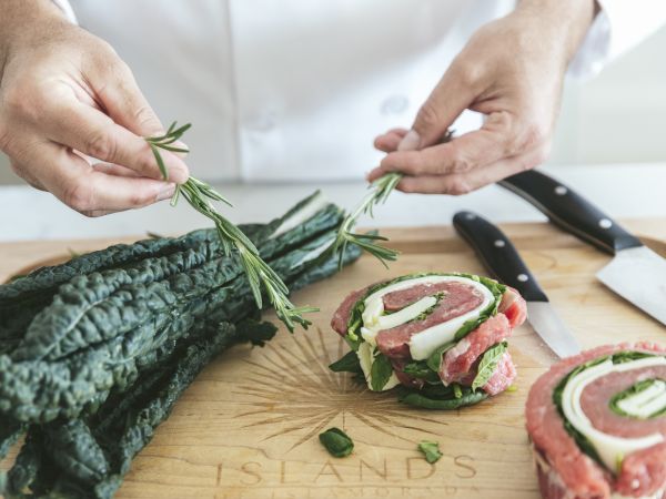 Hands tie rosemary and herbs around a rolled meat preparation on a wooden cutting board with kale nearby.
