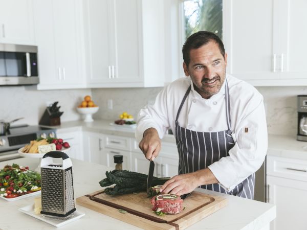 A chef in a bright kitchen chops meat on a wooden board while a box grater and fresh ingredients sit nearby.