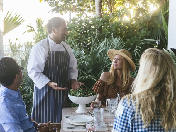 A chef in an apron serves a salad to a small group of diners outdoors at a table surrounded by greenery.