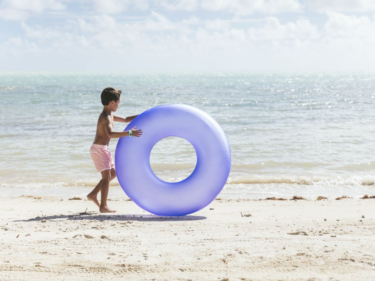 A boy on a sunny beach carries a purple inflatable ring while near the ocean.