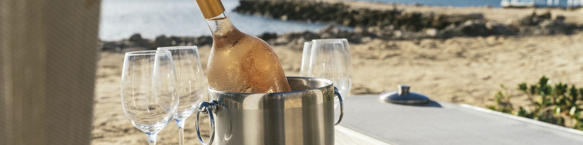 A champagne bottle in an ice bucket with two glasses on a beach table, overlooking the ocean.