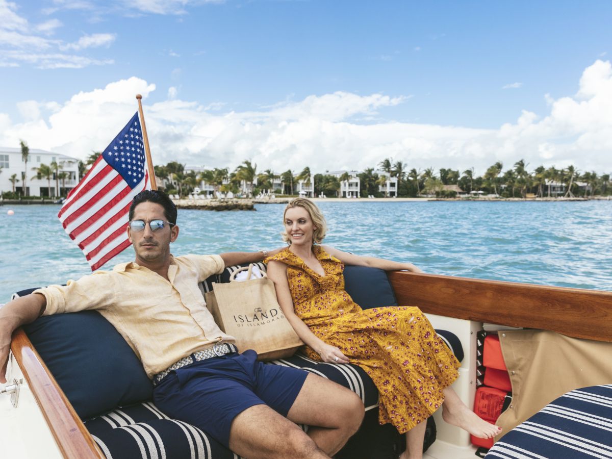 A couple relaxes on a boat by the ocean with an American flag flying as sunny clouds and palm resorts fill the background.
