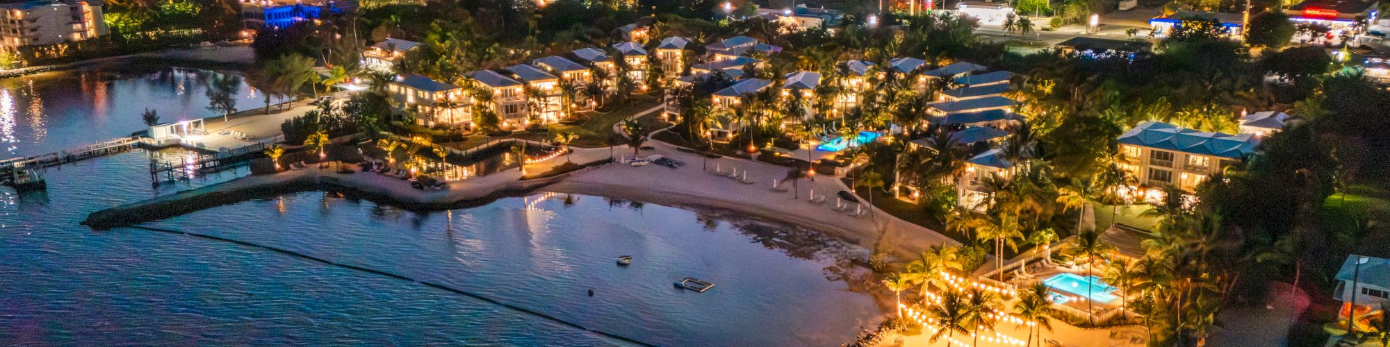 A tropical beach resort at sunset with calm ocean waves, illuminated cottages, palm trees, and a coastline.