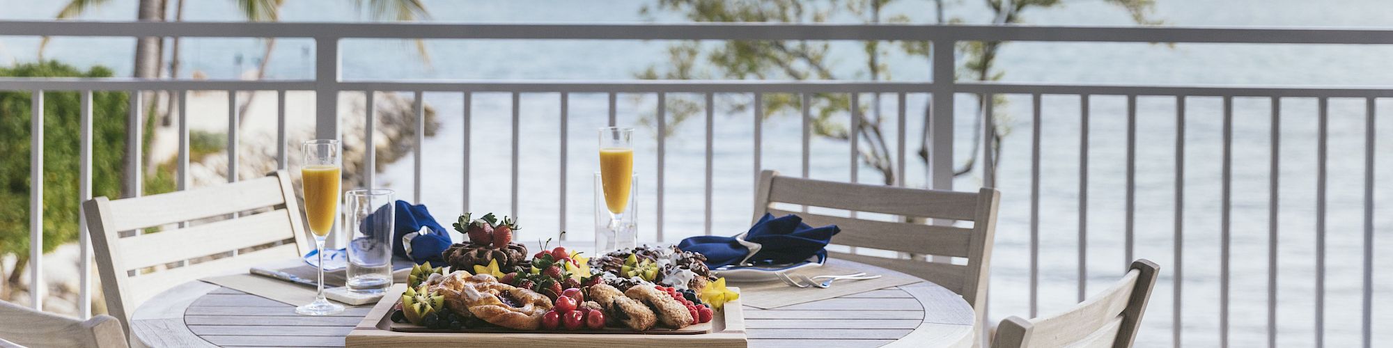 Table set with fruit and drinks on a seaside balcony, with palm trees and the ocean under a cloudy sky.