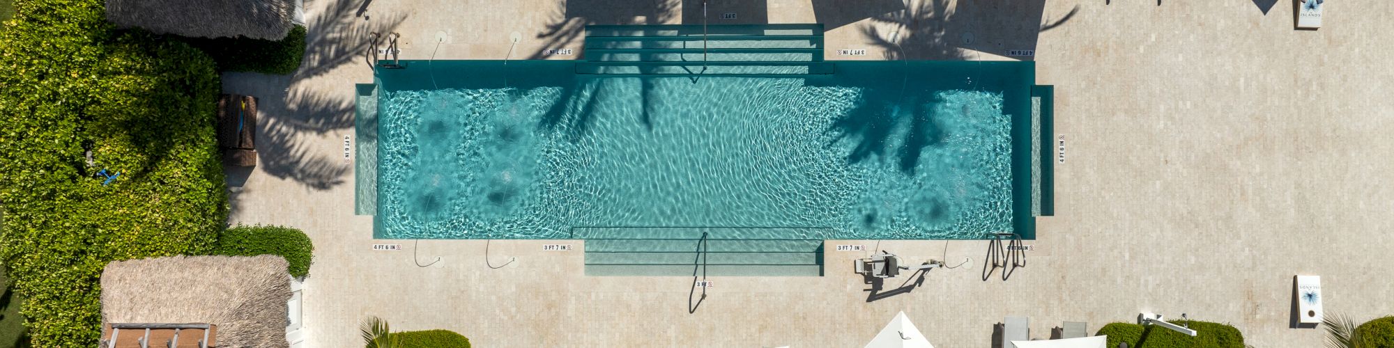 Aerial view of a tropical resort pool surrounded by lush greenery, palm trees, lounge beds, and white umbrellas for sunbathing.