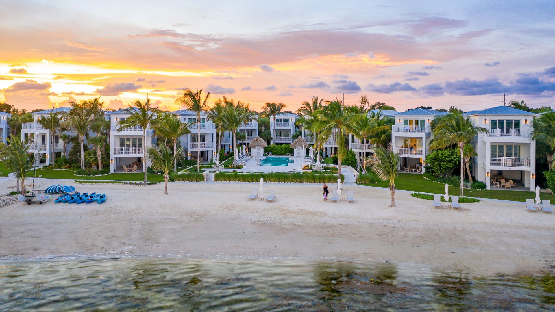 Sunset over a beachfront resort: white villas, palm trees, sandy yard, and a central turquoise pool.