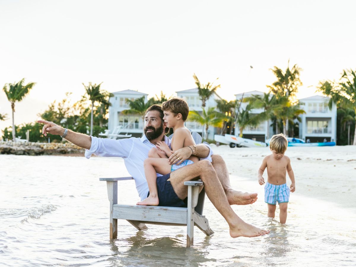 A bearded dad points out something on the beach while holding a child, with another little boy walking nearby.