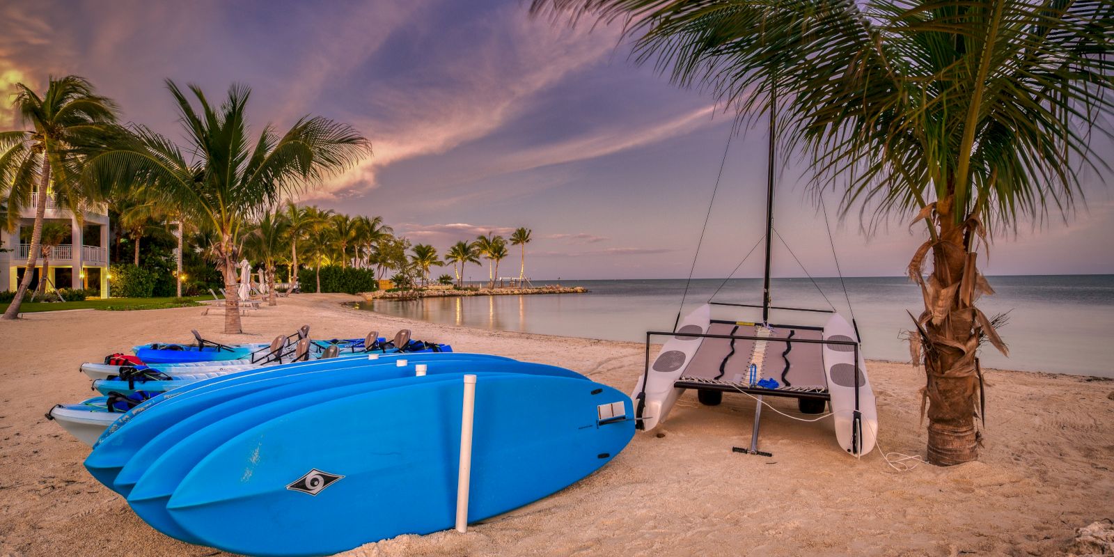 Tropical beach at sunset with blue kayaks, a sailboat, and palm trees beside calm ocean water.
