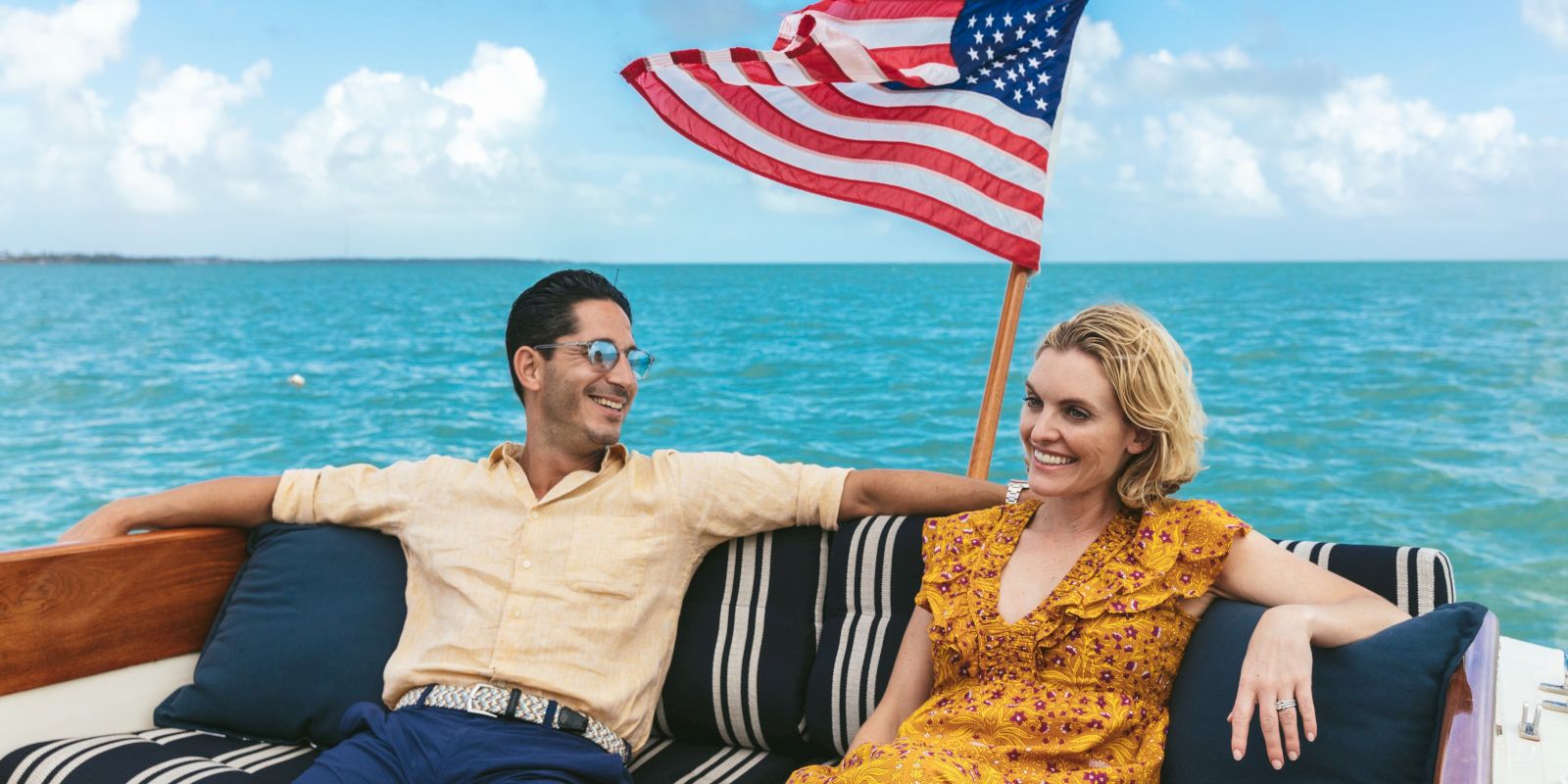 A smiling couple relax on a boat by the ocean, waving a large American flag in the sunshine.