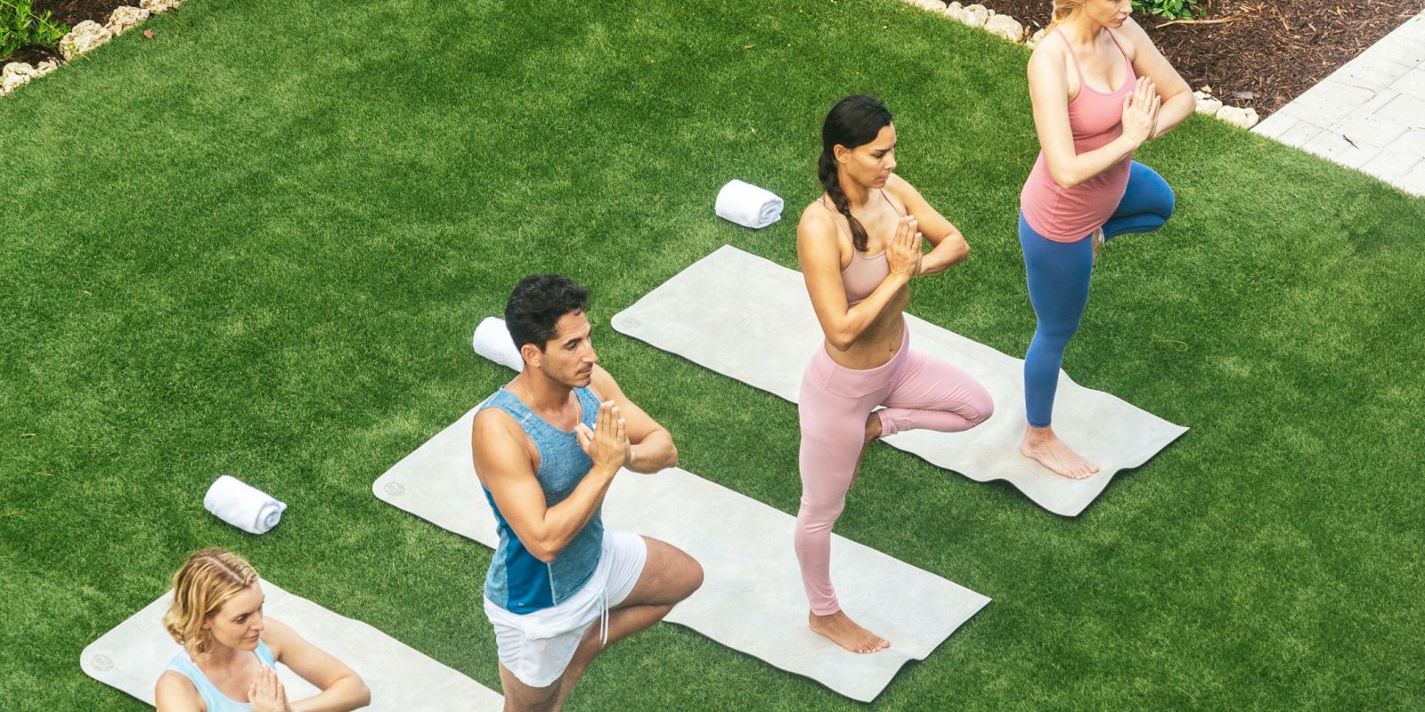 People practice yoga on mats in a sunny outdoor garden, joining a group balancing pose near a swimming pool fence.
