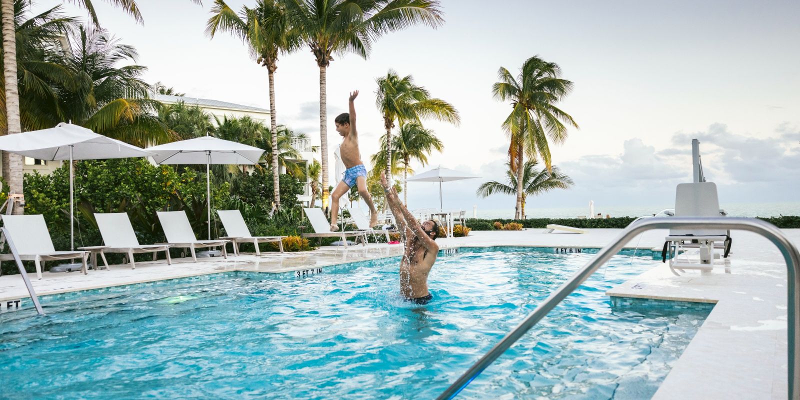 A shirtless man jumps into a sunny resort pool beside palm trees, with lounge chairs and umbrellas in the background.